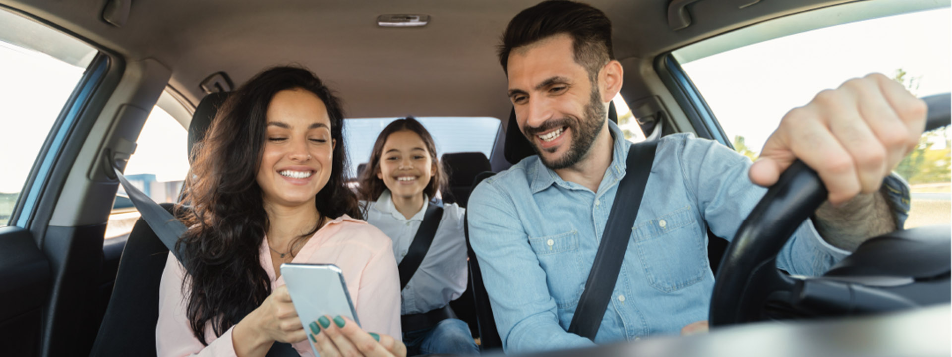 A man driving a car and smiling, with two women passengers; one is looking at a smartphone and the other in the back seat is laughing.