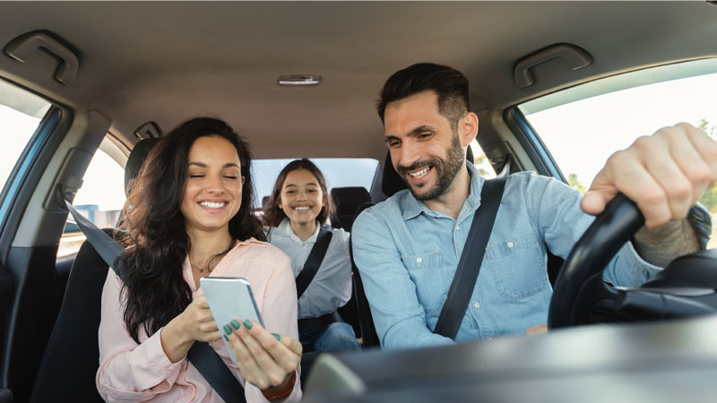 A man driving a car and smiling, with two women passengers; one is looking at a smartphone and the other in the back seat is laughing.