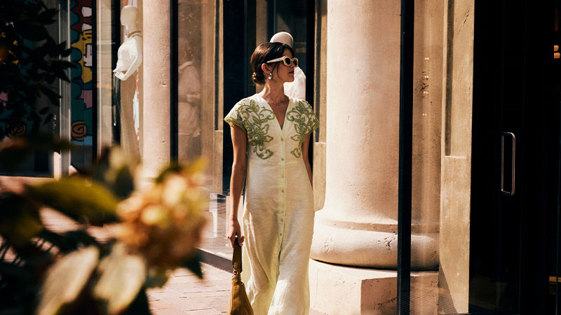 A woman wearing sunglasses and a light dress with green embroidery walks past shop windows and columns on a sunny day.
